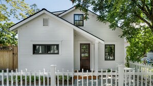 A white two-story house with a front porch, a white picket fence, and a small garden area. - Charming Loft Bedroom Cottage in Downtown White, 1 block away from everything (Whitefish)