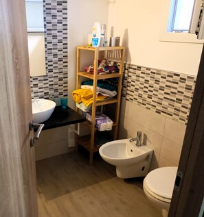 Et badeværelse med vask, toilet og bidét. Der er en hylde med toiletartikler og håndklæder.