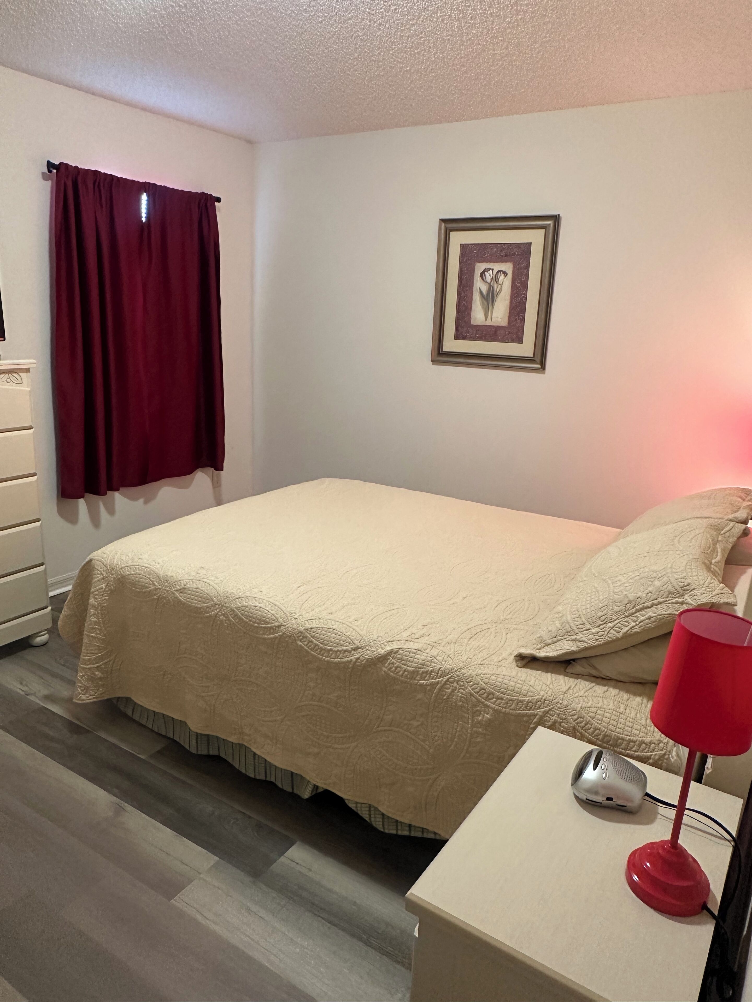 A neatly arranged bedroom with a bed, a bedside table with a lamp, a dresser, and a framed picture on the wall.