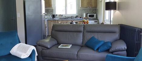 Una sala de estar con un sofá, dos sillones, una mesa de centro con piezas de ajedrez y una cocina al fondo.