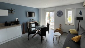 Ein modernes Wohnzimmer mit einem grauen Sofa, einem Esstisch mit Stühlen, einem an der Wand befestigten Fernseher und einem großen Fenster mit Blick nach draußen.