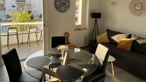 Ein modernes Wohnzimmer mit Glastisch, dunklen Sofas und einem Balkon mit Blick auf Gebäude und einen Baum.