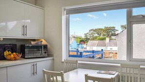 A kitchen with white cabinets, a microwave, and a fruit bowl. There is a window with a view of a blue play structure and houses. - 4 bedroom accommodation in Amlwch (Amlwch)