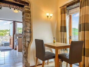 Un comedor con pared de piedra, una mesa de madera, dos sillas, un jarrón con flores y vistas al exterior.