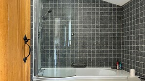 Een moderne badkamer met een douche met glazen wand, een wit bad en donkere betegelde muren.