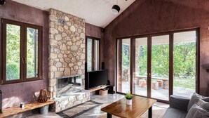 A modern living room with a stone fireplace, large windows, and a flat-screen TV. - Hidden Stone Chalet (Aigialeia)