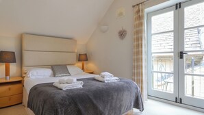 A bedroom with a large bed, two bedside tables with lamps, a grey blanket, and a view of the outside through a glass door. - 3 bedroom accommodation in Somerford Keynes (Somerford Keynes)