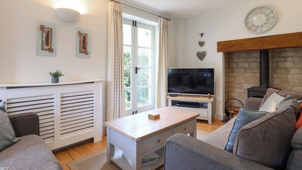 A living room with a sofa, a coffee table, a TV, a fireplace, and a clock on the wall. - 3 bedroom accommodation in Somerford Keynes (Somerford Keynes)