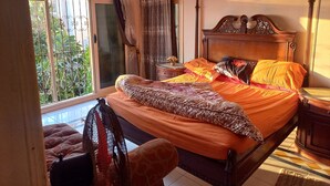 Un dormitorio con una cama grande, cabecera de madera, una mesita de noche, un ventilador y vistas al verdor exterior.