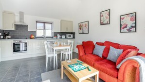 A modern kitchen with white cabinets, a dining area with a wooden table, and a red sofa with cushions. - 1 bedroom accommodation in Hewish (Hewish)