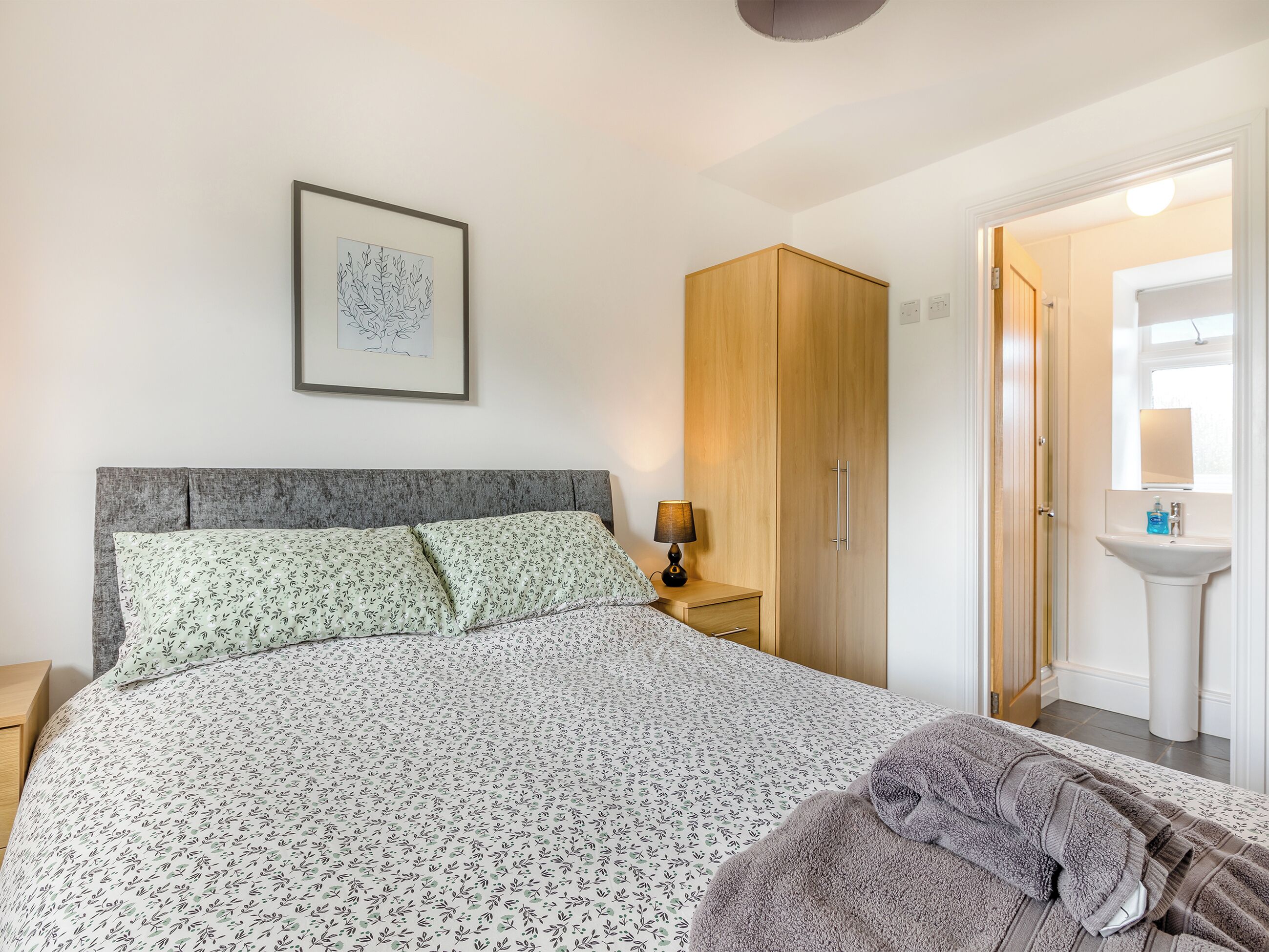 A bedroom with a bed, a nightstand, a wooden wardrobe, a framed artwork, and a bathroom with a sink and mirror.