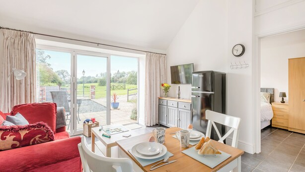 A bright, well-lit room with a dining table set for two, a red sofa, and a kitchen area with a refrigerator and a clock on the wall. - 1 bedroom accommodation in Hewish (Hewish)