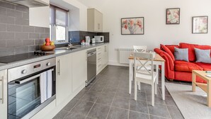 A modern kitchen with white cabinets, a built-in oven, and a dining area with a red sofa and a table. - 1 bedroom accommodation in Hewish (Hewish)