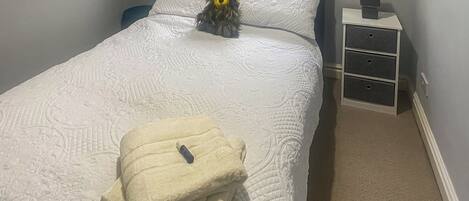 Una cama bien hecha con una colcha blanca y una toalla doblada, una mesita de noche con un altavoz, y una ventana con persianas.