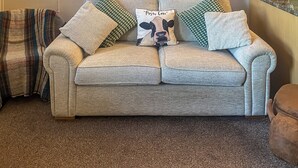 Een beige bank met een kussen in de vorm van een koe, groene kussens met een patroon en een geruit plaid.