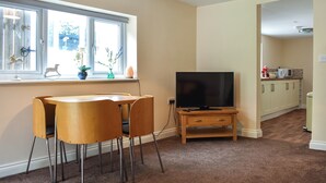 Een woonkamer met een televisie op een standaard, een eettafel met stoelen, een raam met planten, en een keuken op de achtergrond.