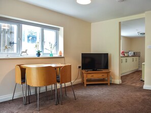 Ein Wohnzimmer mit Fernseher auf einem Schrank, einem Esstisch mit Stühlen, einem Fenster mit Pflanzen und einer Küche im Hintergrund.