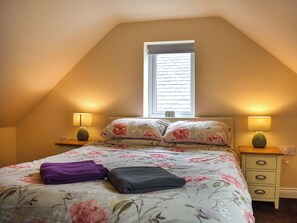 Un dormitorio con techo inclinado, una ventana con persianas, dos lámparas de noche, una cama con ropa de cama floral, una toalla morada y una almohada gris.