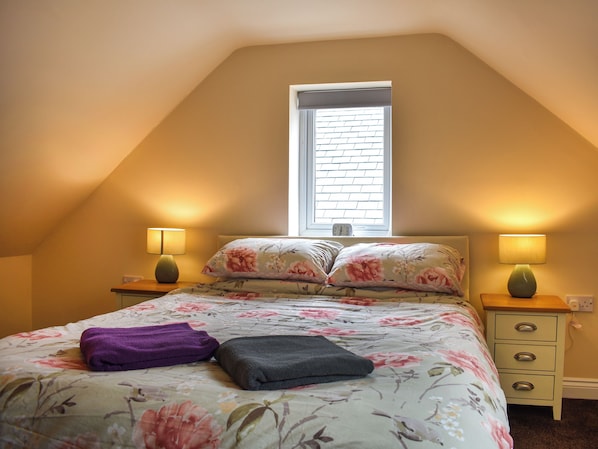 Un dormitorio con techo inclinado, una ventana con persianas, dos lámparas de noche, una cama con ropa de cama floral, una toalla morada y una almohada gris.