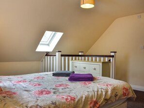 Ein Schlafzimmer mit schrägem Dach, einem Oberlicht, einem Bett mit floralem Bettbezug, einem Nachttisch und einer Kommode mit Schubladen.