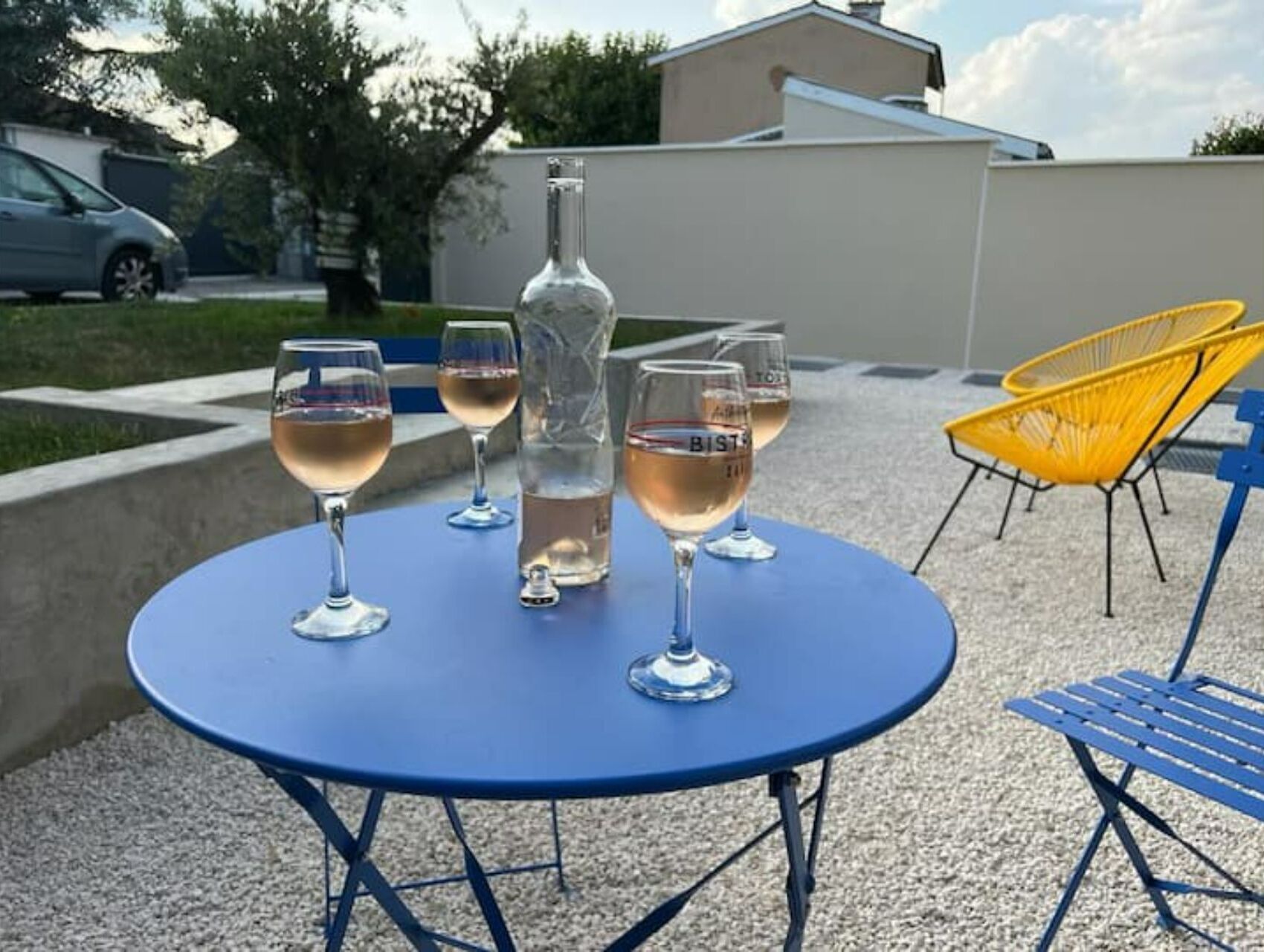 Una mesa azul al aire libre con cuatro copas de vino y una botella, rodeada de sillas y un edificio al fondo.