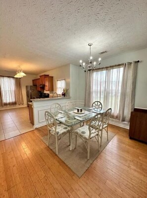 A spacious dining area with a glass table and white chairs, a kitchen in the background, and a chandelier. - Sleeps 8: King sized bed - Home away from home, yet close to everything! (Douglasville)