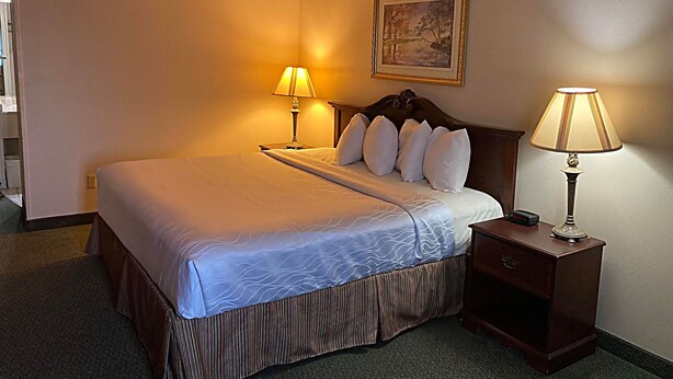 A hotel room with a bed, two bedside lamps, a nightstand, a painting on the wall, and a carpeted floor. - The Jeffreys Hotel Extended Stay (Osceola)