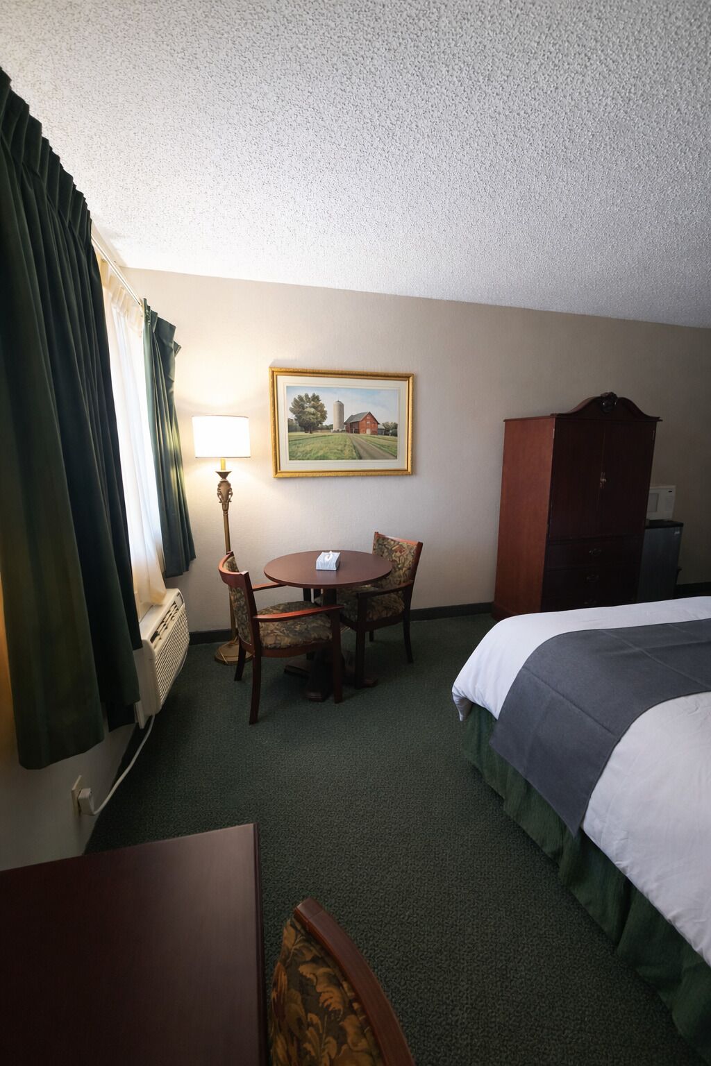 A hotel room with a bed, a desk with a lamp, a chair, a painting on the wall, and a wardrobe.