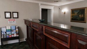 Un área de recepción con mostrador de madera, certificados enmarcados, un letrero de advertencia, un cuadro enmarcado y un tablón de anuncios con folletos de viajes.