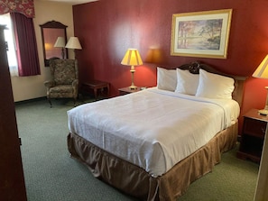 A hotel room with a large bed, a chair, a small table, a lamp, and a painting on the wall.