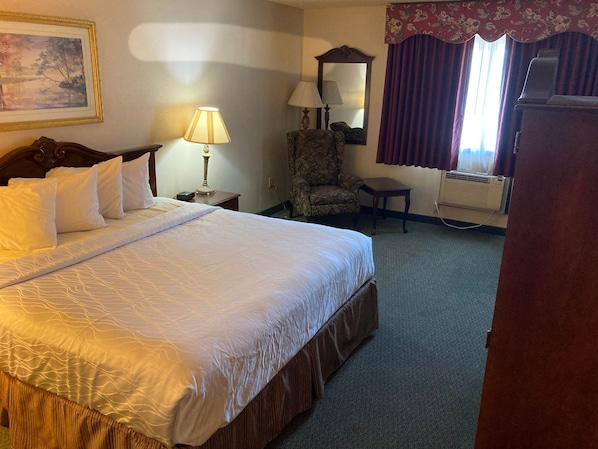 A hotel room with a large bed, two bedside lamps, a chair, a small table, a mirror, and an air conditioning unit.