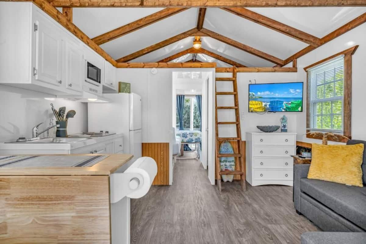 Een compacte, moderne keuken met witte kasten, een ingebouwde magnetron en een houten aanrecht. Een ladder leunt tegen de muur, en boven een witte dressoir hangt een televisie.