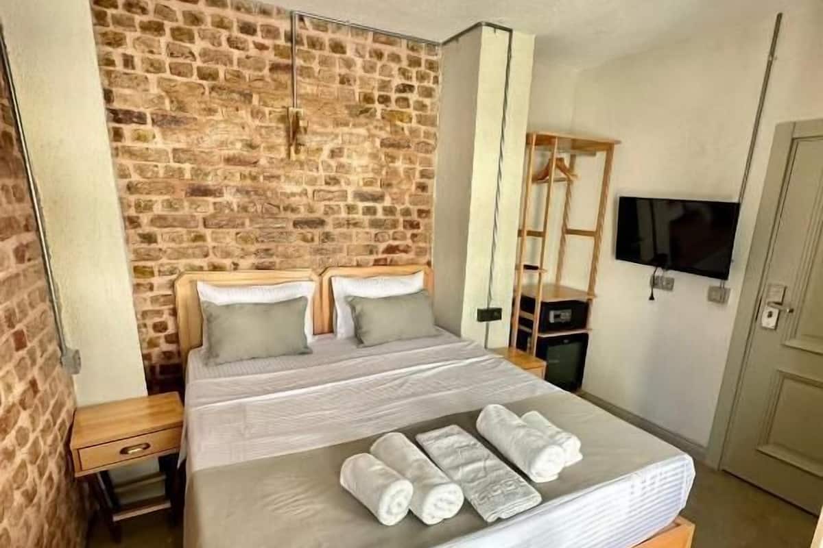 A bedroom with a brick wall, a bed with rolled towels, a wooden nightstand, a TV, and a mirror.