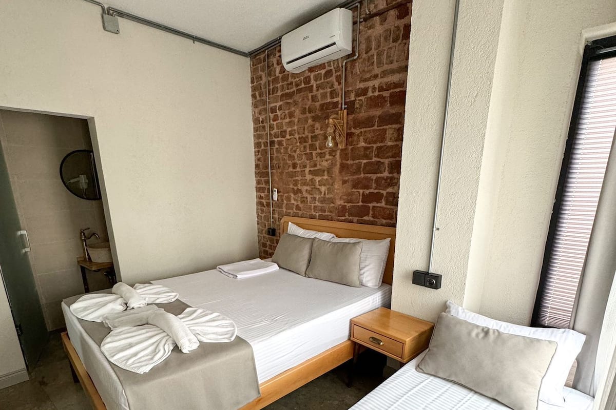 A hotel room with a brick accent wall, a large bed with white linens, a smaller bed, a wooden nightstand, and an air conditioning unit.