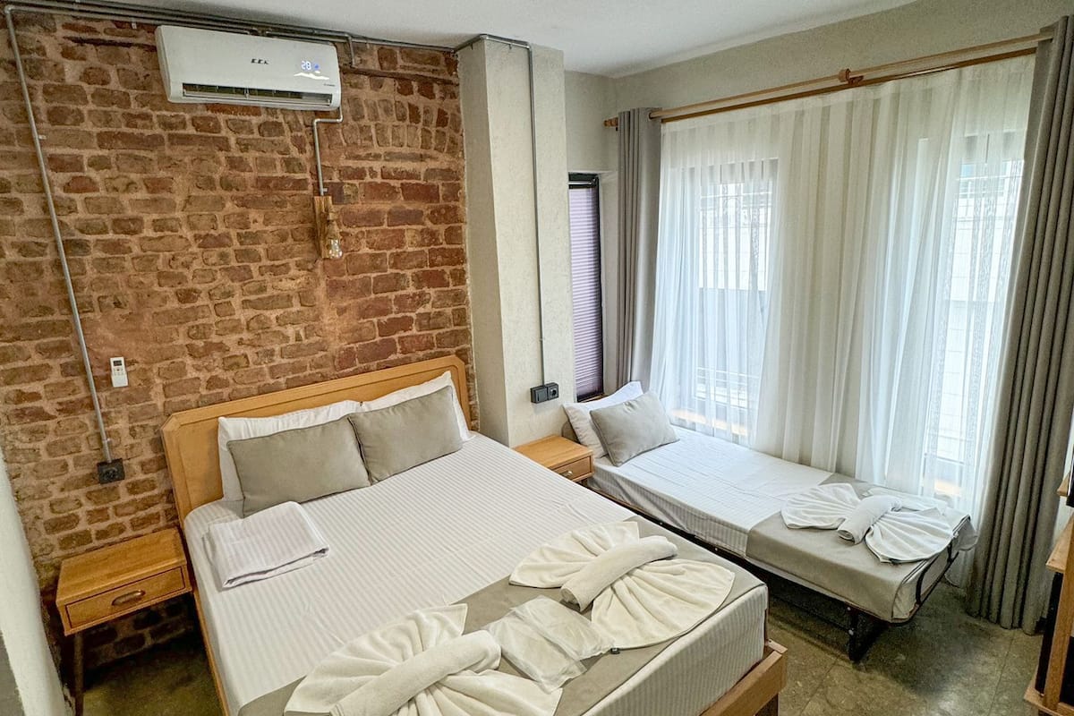 A hotel room with a brick wall, a large bed with towels, a smaller bed, a window with curtains, and an air conditioning unit.