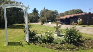 Un jardín bien cuidado con una pérgola blanca, una fuente decorativa y un edificio de techo rojo al fondo.