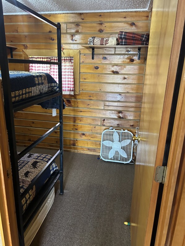 A bunk bed room with a wooden wall, a small shelf with blankets, and a white fan.
