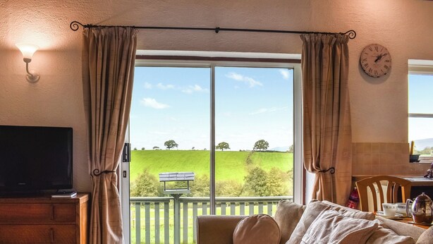 A living room with a sofa, a television, a clock, and a view of a green field through a large window. - 1 bedroom accommodation in Tatham, near Lancaster (Tatham, near Lancaster)