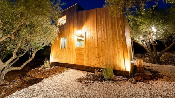 A wooden cabin with a single entrance and two windows, surrounded by trees and gravel.