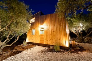 Una cabaña de madera con una sola entrada y dos ventanas, rodeada de árboles y grava.