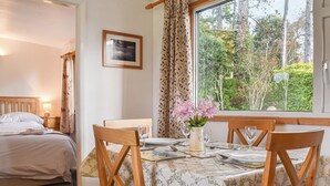 Un coin repas bien aménagé, avec une table dressée pour deux personnes, un lit doté d’une tête de lit en bois et une fenêtre agrémentée de rideaux.