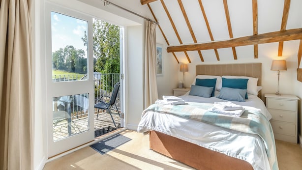 Een slaapkamer met een grote schuifpui naar een buitenruimte, een bed met veel kussens, een nachtkastje met een lamp en een houten plafondconstructie.