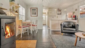 A cozy living room with a fireplace, a dining area with a table and chairs, a black leather armchair, and a kitchen area with cabinets and appliances. - Nice home in Sydals with WiFi (Sydals)