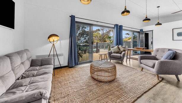 Een moderne woonkamer met een grijze bank, een rond rieten bijzettafeltje en een grijze fauteuil. Er zijn hanglampen, een vloerlamp en een groot raam met gordijnen.