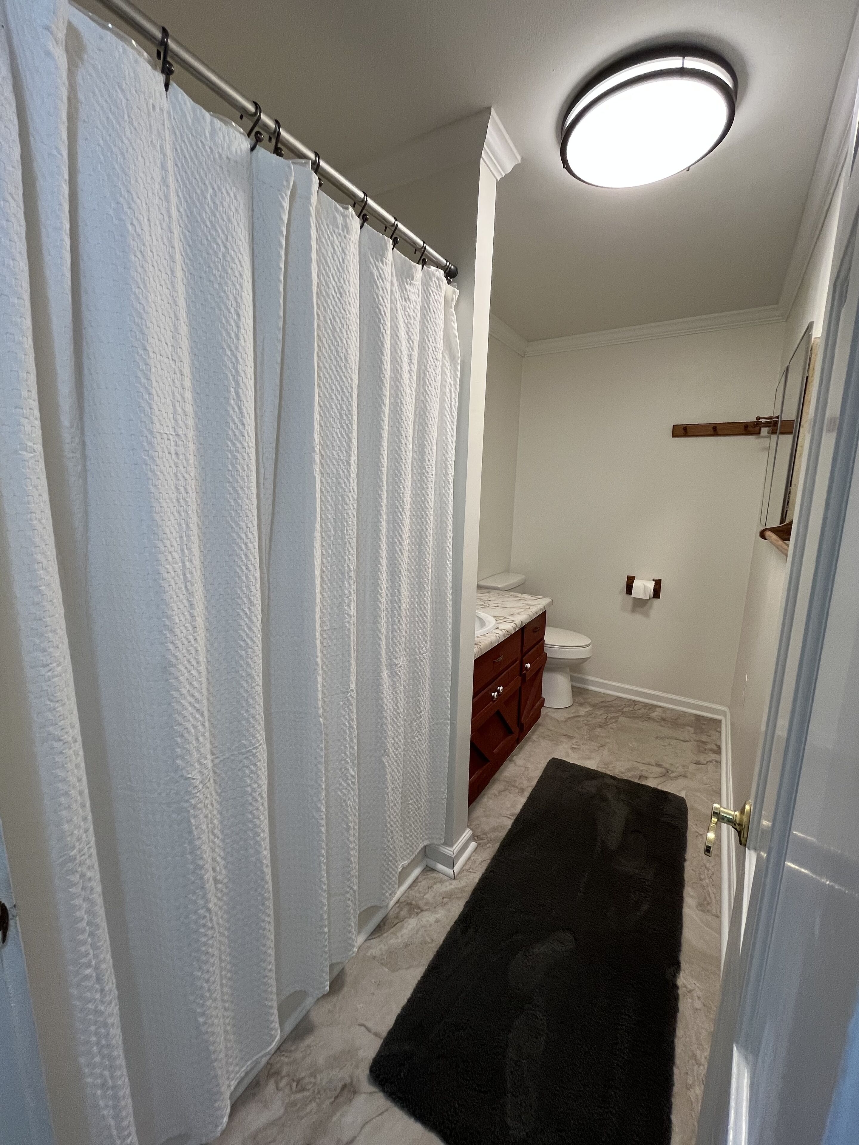 A bathroom with a shower curtain, a toilet, a vanity with a mirror, and a black rug on the floor.