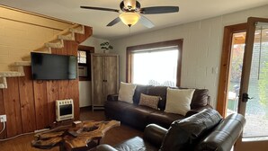 A living room with a brown leather sofa, a wooden coffee table, a TV mounted on a wooden wall, a ceiling fan, and a window with blinds. - Stenson Hollow Lake Condo (Muscle Shoals)