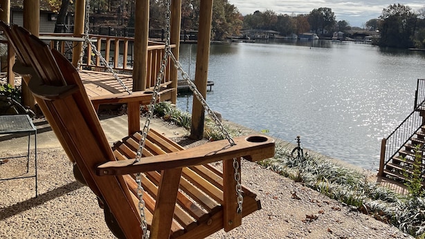 A wooden swing chair under a covered area overlooking a lake with a dock and trees in the background. - Stenson Hollow Lake Condo (Muscle Shoals)