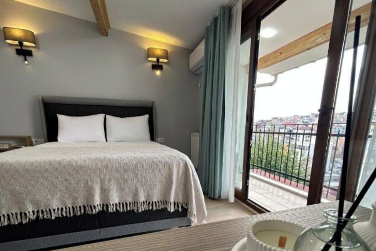 A hotel room with a bed, a round table with a bottle of perfume, and a view of the city through large windows.
