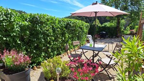 An outdoor patio with a table and chairs, surrounded by greenery and a large umbrella. - Gîte Philomène Dans Ancien Presbytère - 2 Pers - à St Georges de Lévéjac (Massegros Causses Gorges)