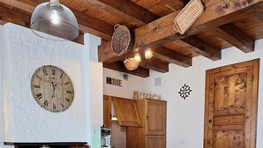 A cozy living room with a fireplace, wooden ceiling beams, a clock, and a dining area with a table set for two. - Gîte Philomène Dans Ancien Presbytère - 2 Pers - à St Georges de Lévéjac (Massegros Causses Gorges)
